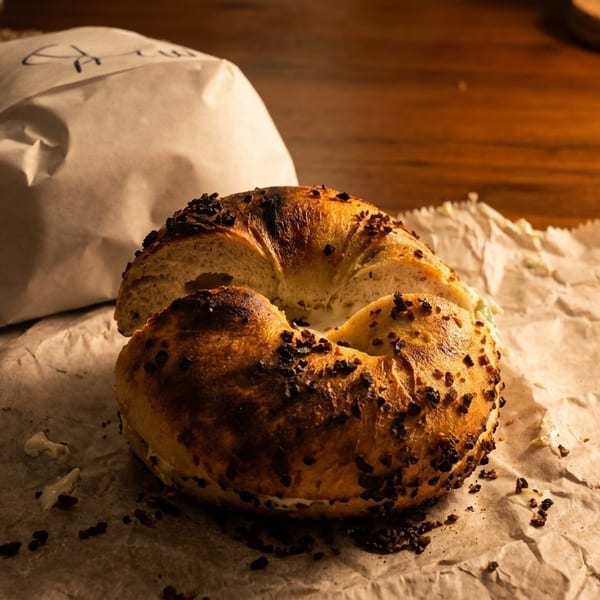 A toasted bagel with cream cheese and seasoning sits on crumpled parchment paper next to a wrapped bagel.