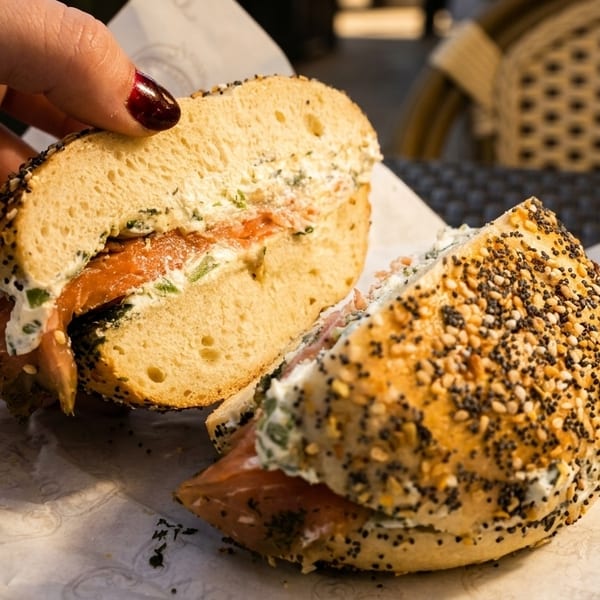 Hand holding a sliced everything bagel with lox and cream cheese on parchment paper.